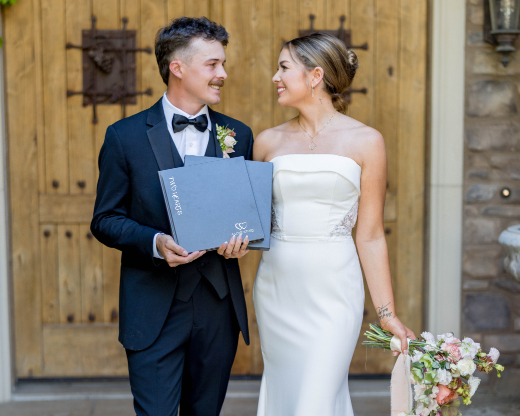 A bride in a strapless white mermaid-style wedding gown and a groom in a black tuxedo with a black bow tie stand in front of a rustic wooden door. The bride holds a bouquet of pink and white flowers, and the groom holds a gray keepsake box.