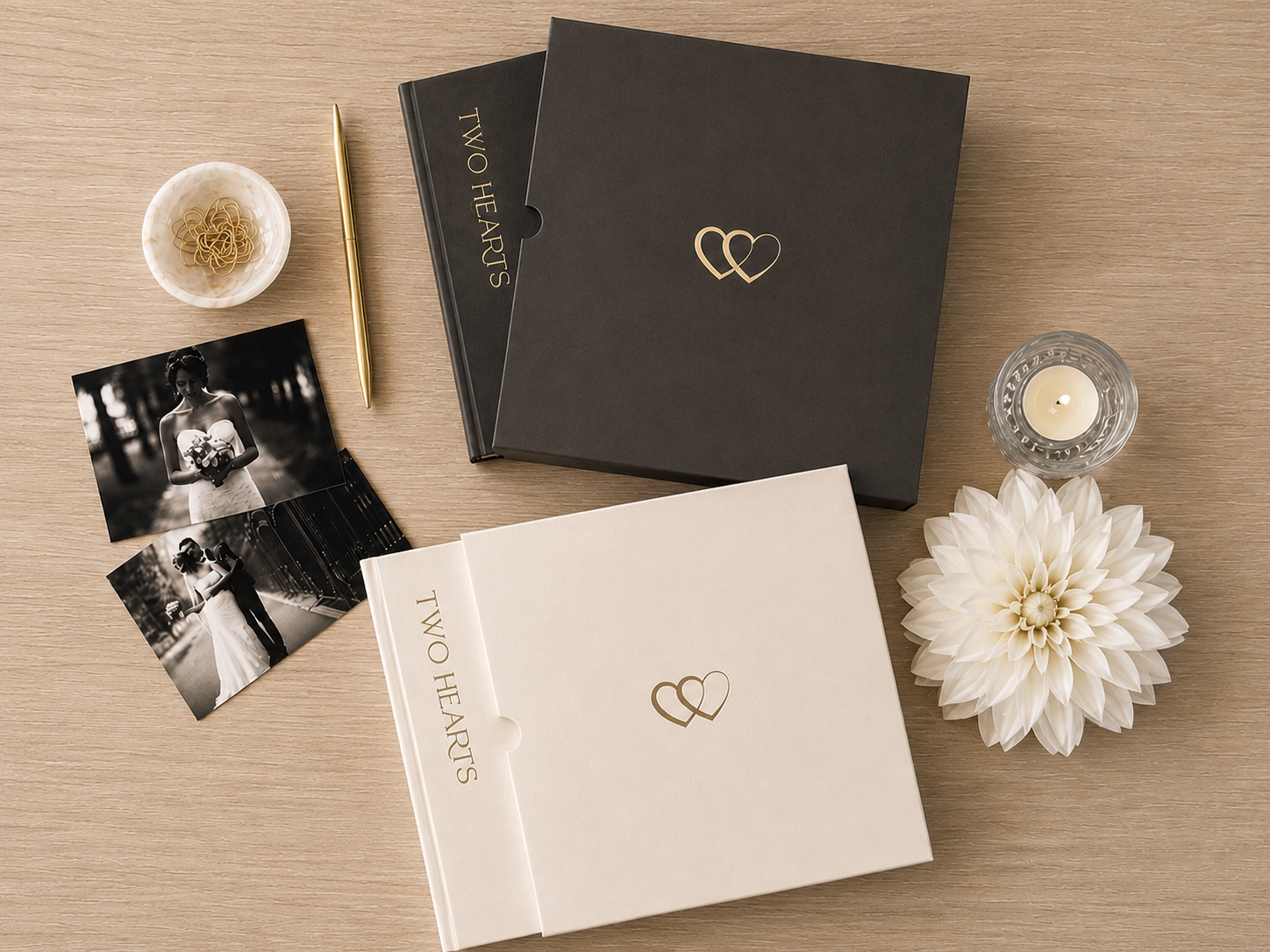 A styled flat-lay of wedding keepsake albums and boxes on a light wood surface, including a black box with a gold double-heart logo, a cream album with a matching logo, printed photos, a candle, and decorative flowers.