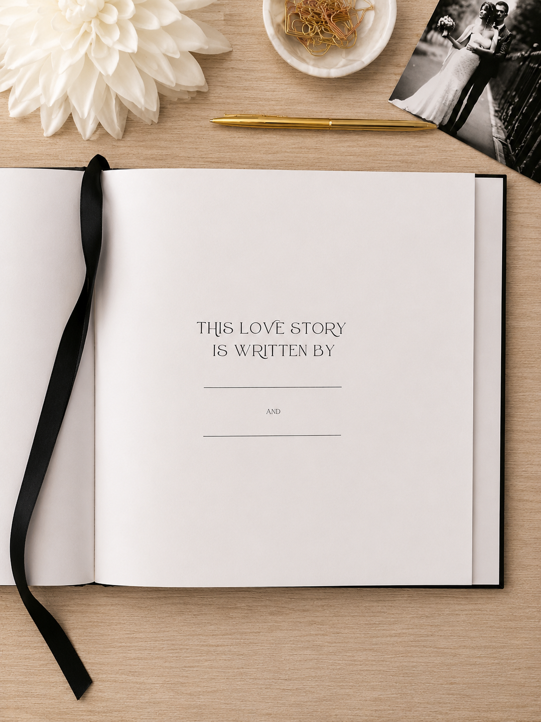 An open wedding guest book or keepsake album on a light wood tabletop, with a black ribbon bookmark draped along the left page. The center page reads, 'THIS LOVE STORY IS WRITTEN BY' with blank lines and 'A&O' below.