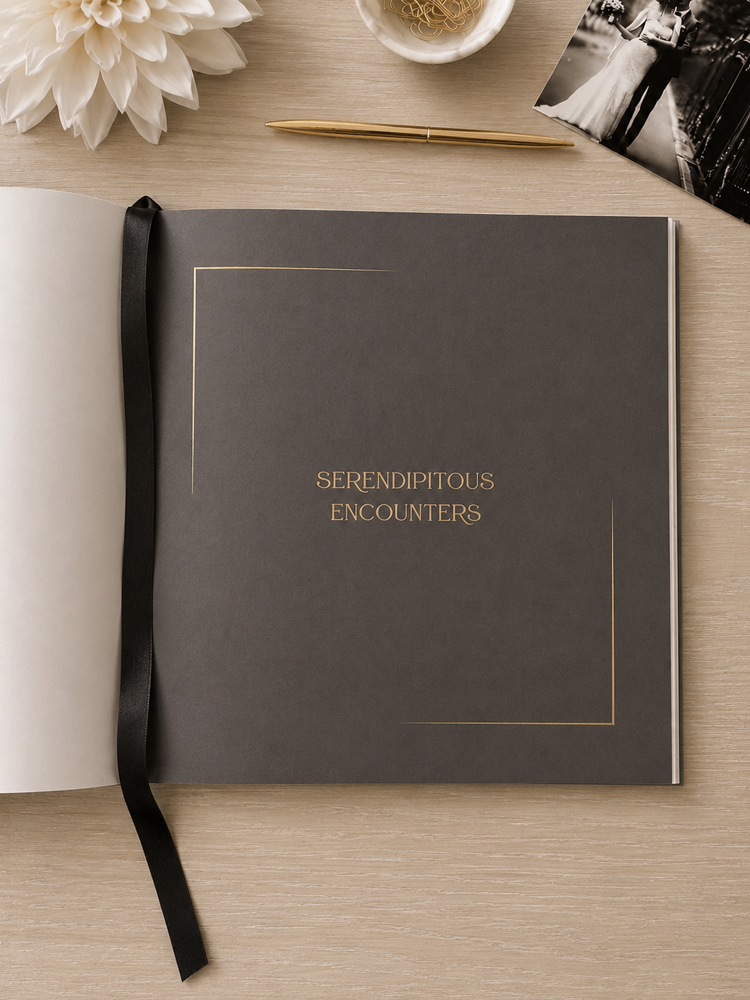 A dark gray wedding guest book with gold foil lettering reading 'SERENDIPITOUS ENCOUNTERS' and a black ribbon bookmark, styled on a light wood tabletop with a white flower, pen, and photo print.