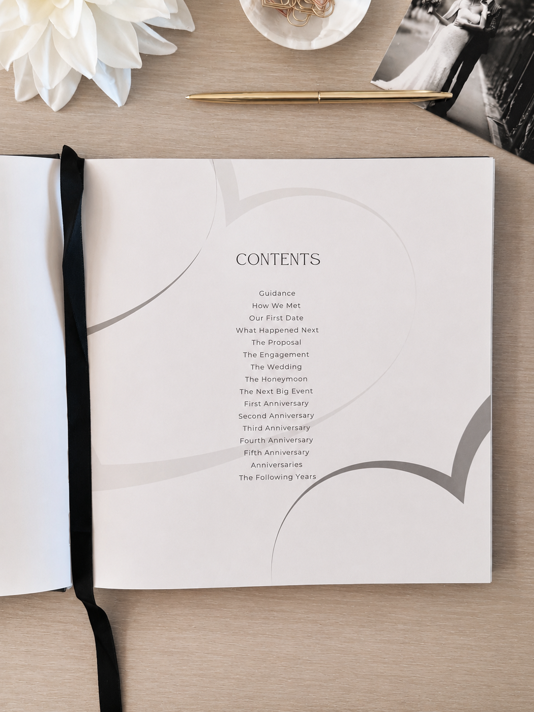An open wedding program or guest book on a light wood tabletop, showing a printed contents page with elegant curved gray line art and a black ribbon bookmark. Decorative props including a white flower, a pen, and a black-and-white photo print are arranged around it.