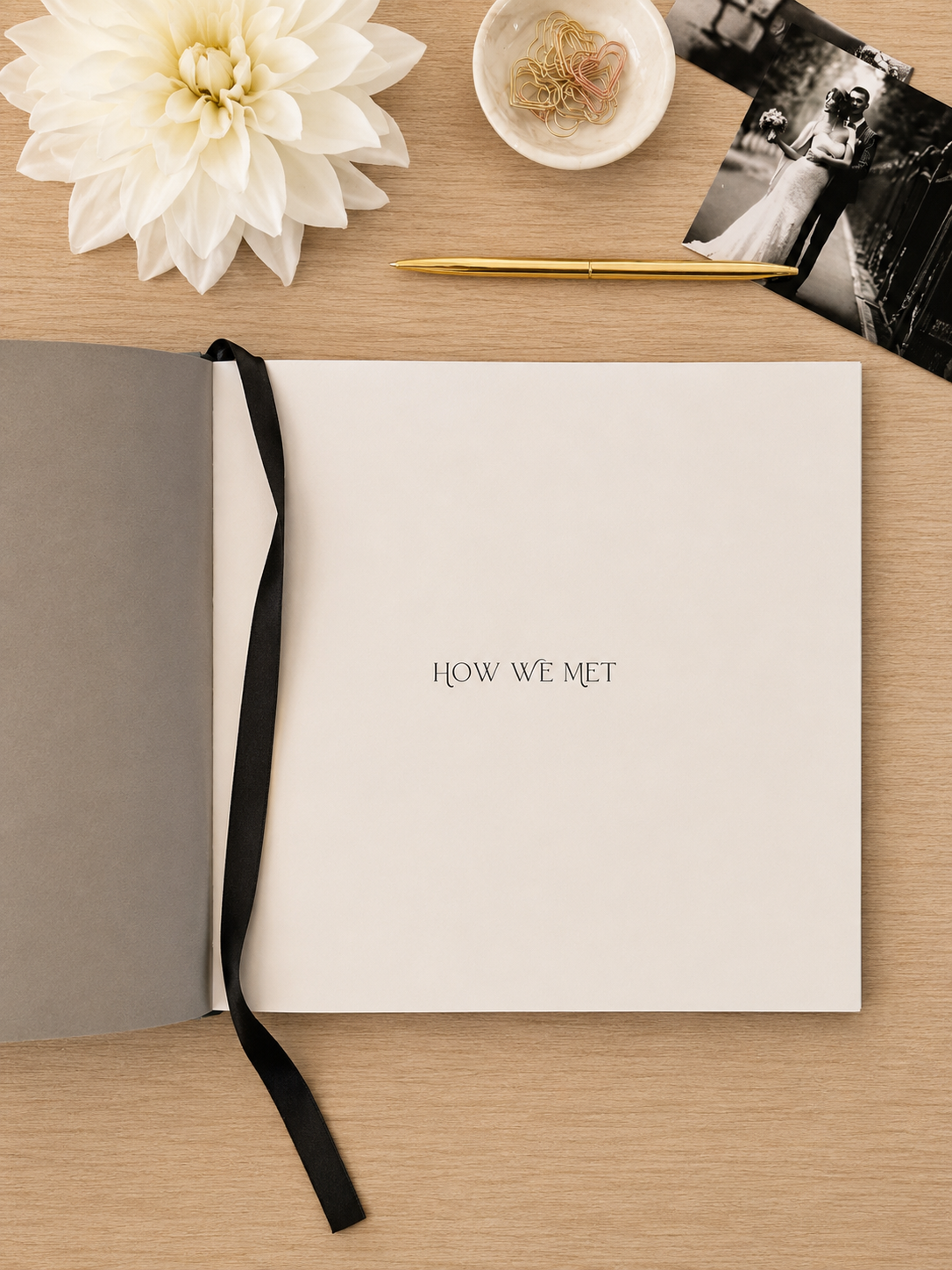 An open stationery keepsake book or guest book on a light wood surface, with the center page reading 'HOW WE MET' in small serif text. A white flower, a small dish, a black-and-white photo, and a gold pen are arranged around it as styled props.