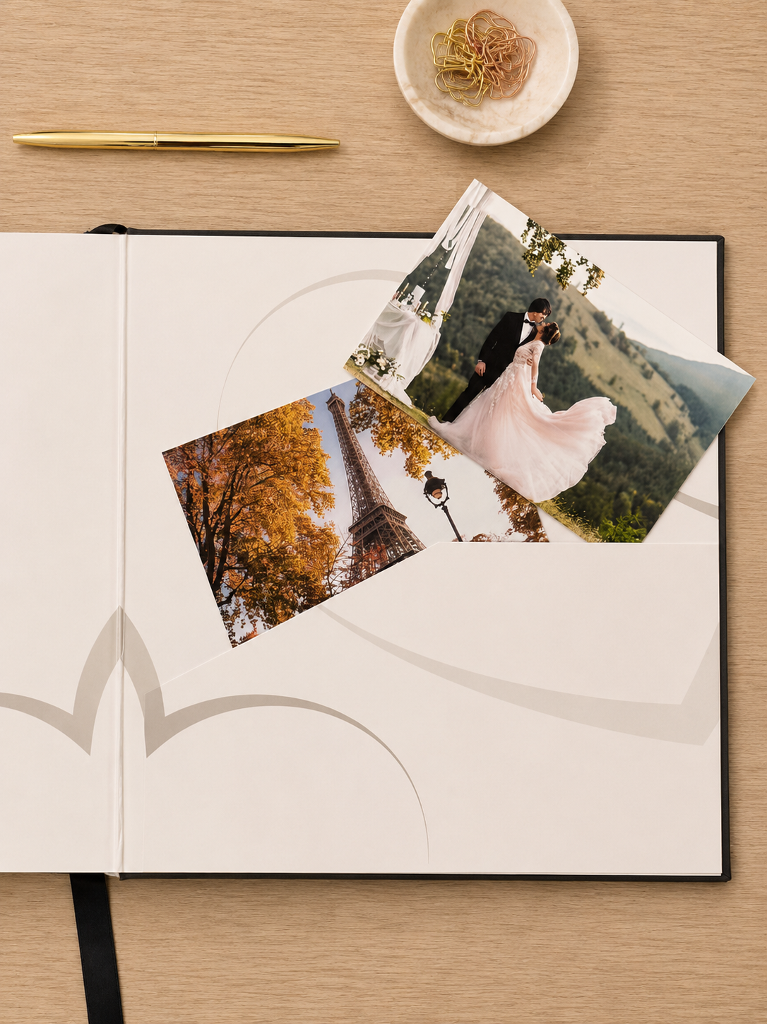 A luxury hardcover photo album spread open on a light wood desk, featuring elegant minimalist page designs and two custom printed wedding photographs placed on top, alongside a gold pen and a small dish of gold binder clips.