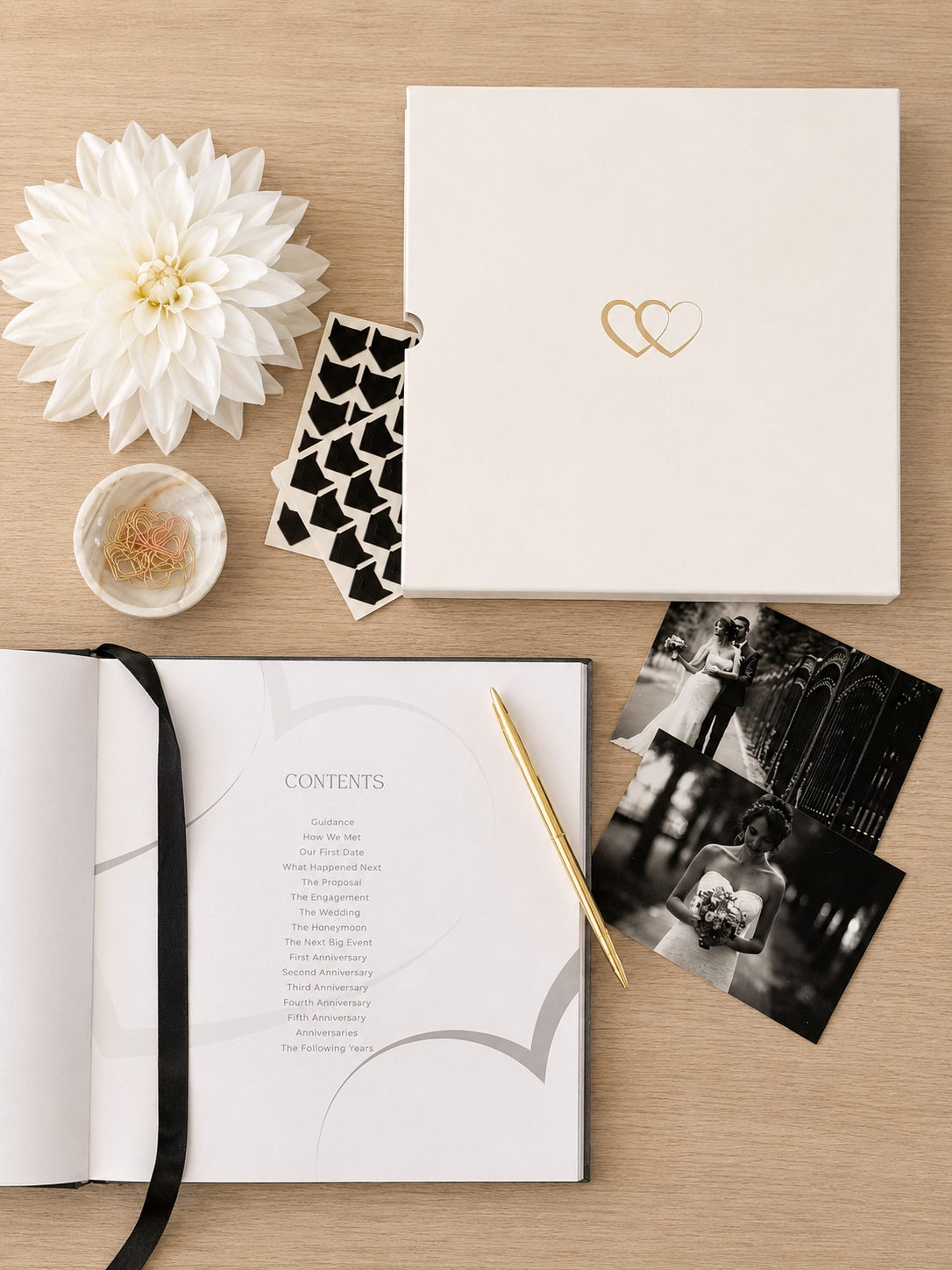 A styled flat-lay of a wedding keepsake album set on a light wood background, featuring an open book with printed contents, black-and-white wedding photos, a white flower, a small dish, a patterned ribbon, and a closed cream-colored album with a gold double-heart logo.